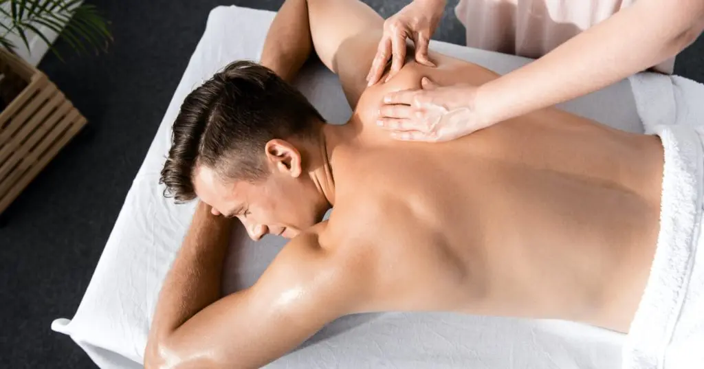 Đôi bàn tay nam giới đang nhẹ nhàng massage vùng lưng cho nữ giới bằng tinh dầu, mở đầu cho liệu trình massage kích thích khoái cảm tại không gian ấm cúng
