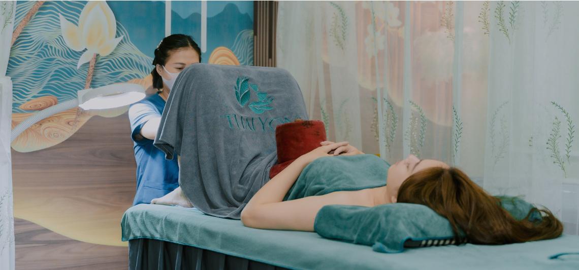 Kỹ thuật viên massage yoni đeo găng tay y tế, sử dụng dầu massage hữu cơ trong phòng trị liệu vô trùng với ga trải giường trắng tinh khôi tại Hà Nội