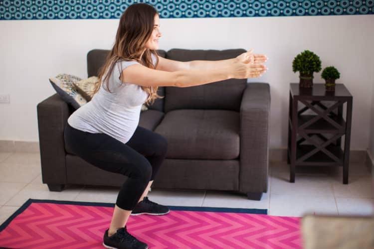 Một phụ nữ mang thai đang thực hiện bài tập thở cơ hoành và squat nhẹ nhàng trong phòng khách sáng sủa, kết hợp với thảm tập yoga.